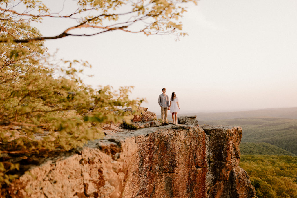 man and woman holding hands on cliff and looking out at sunset