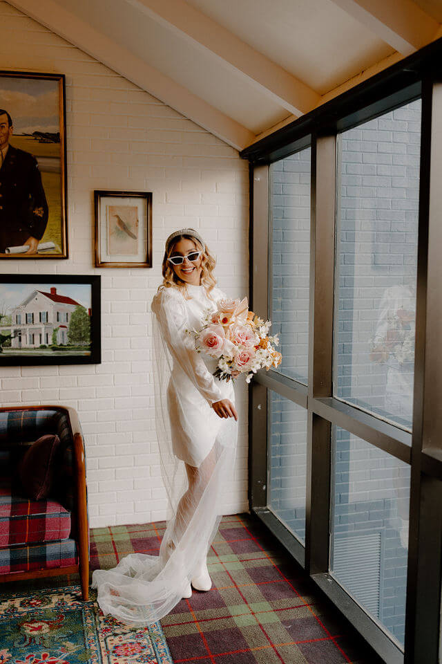 bride in vintage wedding dress and sunglasses smiling while inside building with gallery wall 