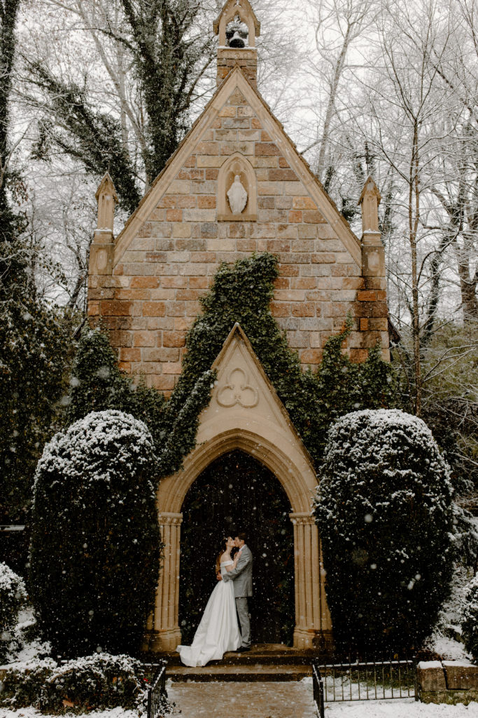 Bride and groom kissing with small chapel in background while it snows -- arkansas elopement photographer